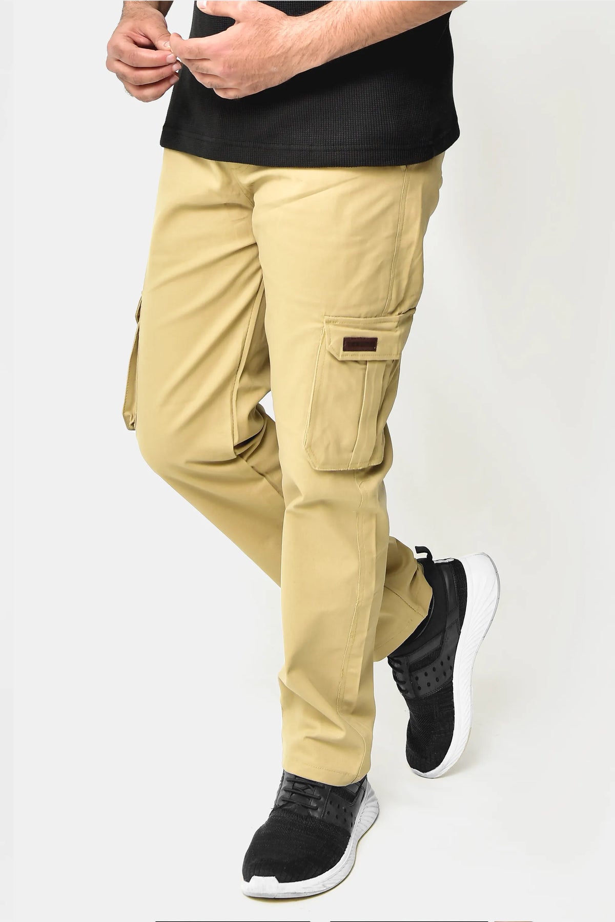 A person standing against a white background, wearing beige cargo trousers with multiple pockets and black shoes.