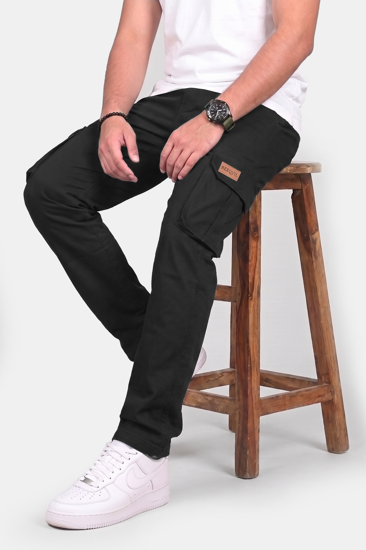 A man wearing black cargo pants sits on a wooden stool, paired with a white t-shirt and white sneakers. The pants feature a brown leather patch on the cargo pocket.