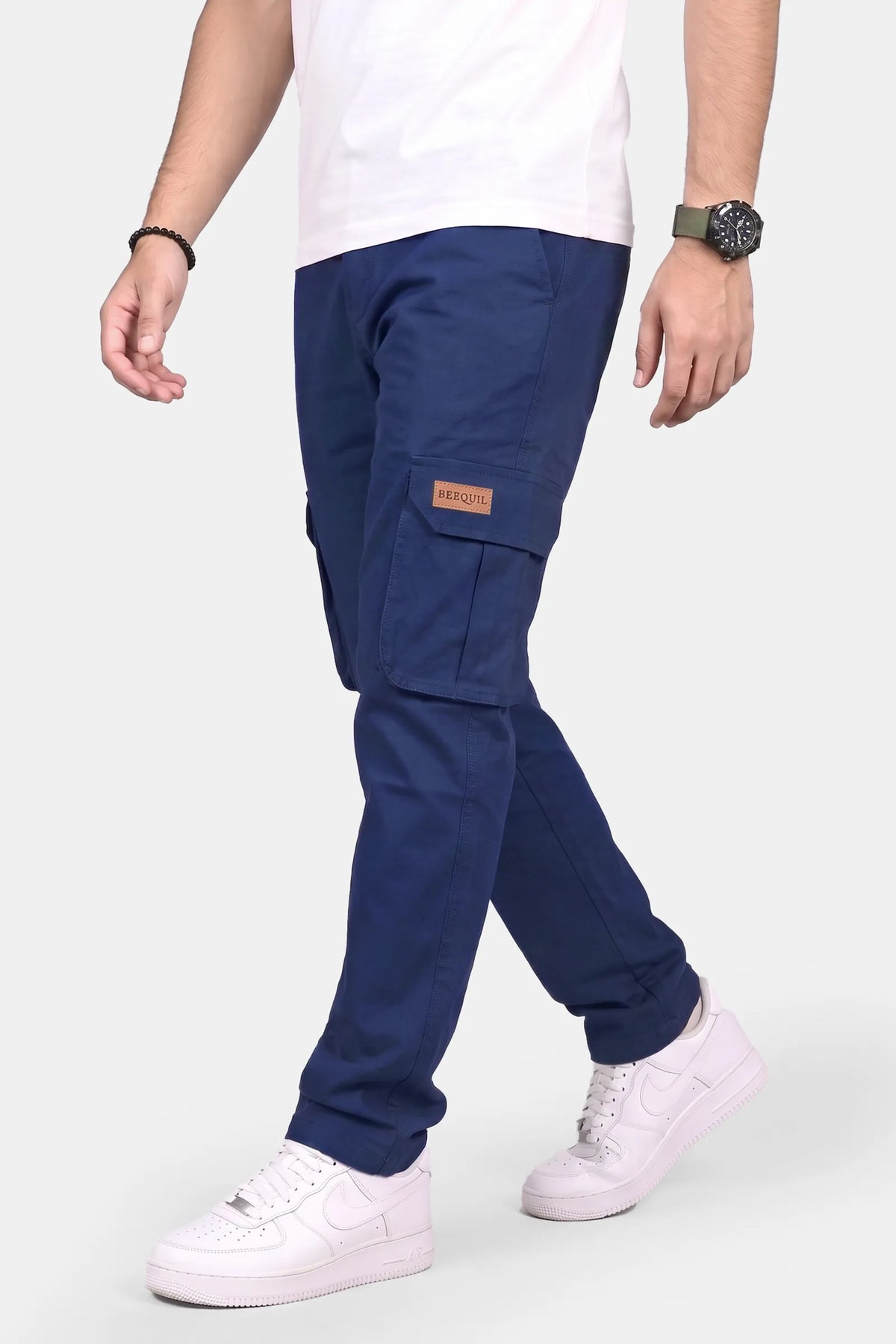 A person wearing navy blue cargo pants with a small brown brand patch on the side pocket, paired with a white t-shirt and white sneakers, shown from the waist down against a light gray background.