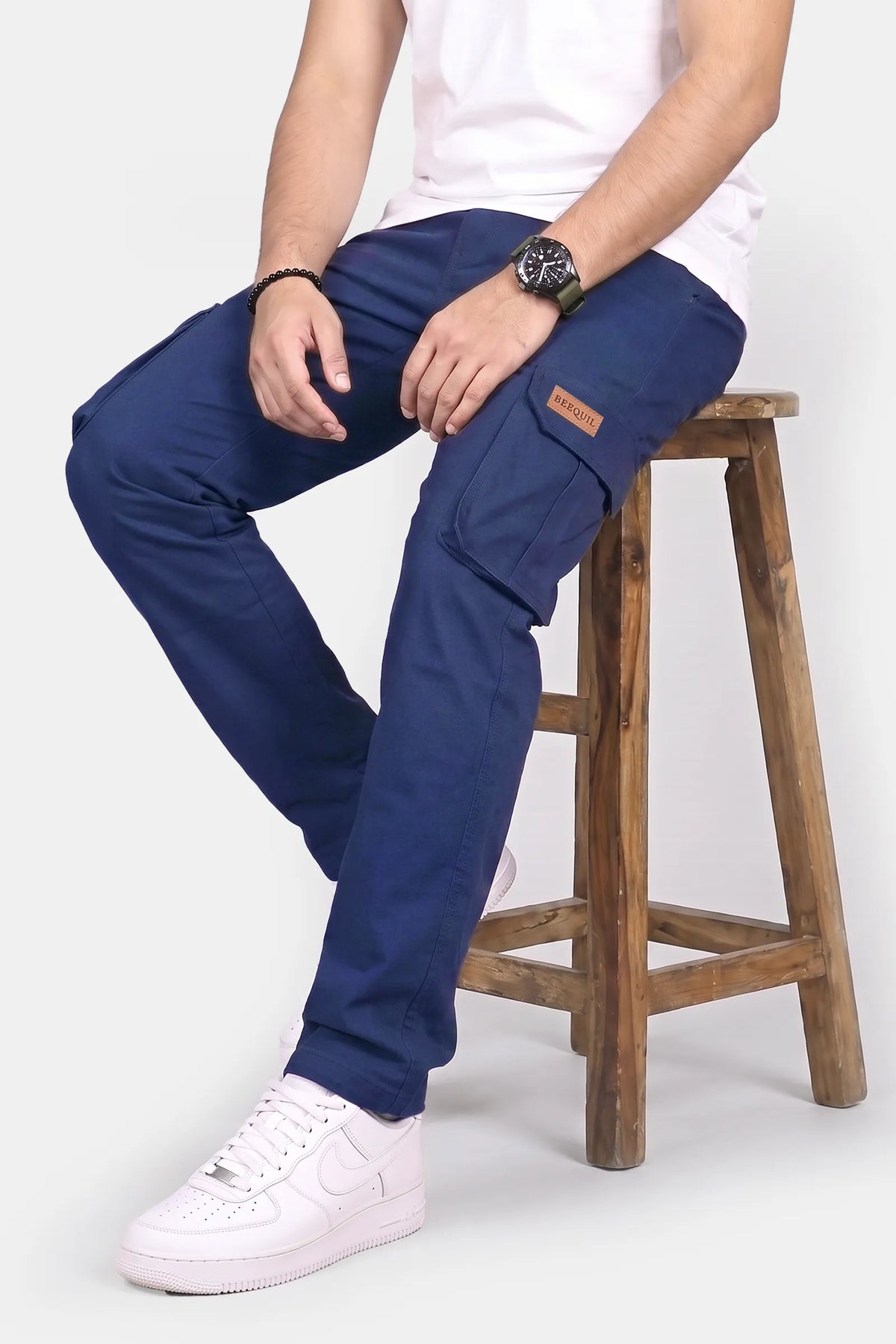 A person seated on a wooden stool wearing navy blue cargo pants, a white T-shirt, and white sneakers against a light gray studio background.