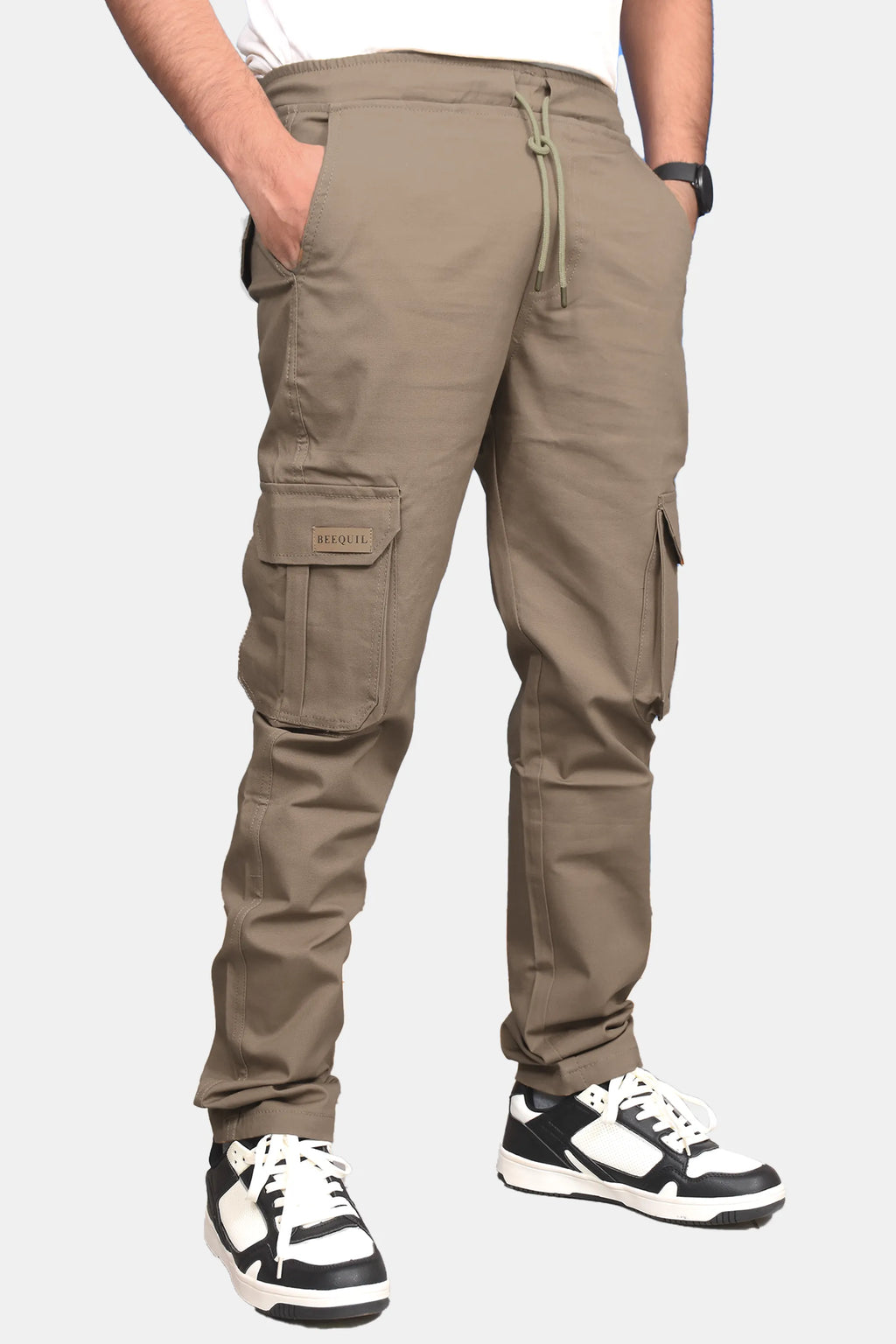 A person wearing khaki cargo jogger pants with side flap pockets and a drawstring waist, paired with black and white sneakers, shown from the waist down on a light gray background.