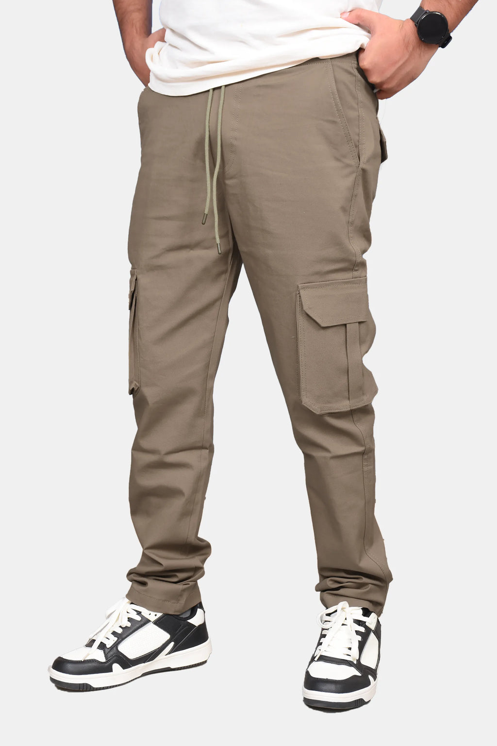 A person wearing taupe-brown cargo jogger pants with side flap pockets, a drawstring waist, and elastic cuffs, paired with a white t-shirt and black-and-white sneakers against a light gray background.