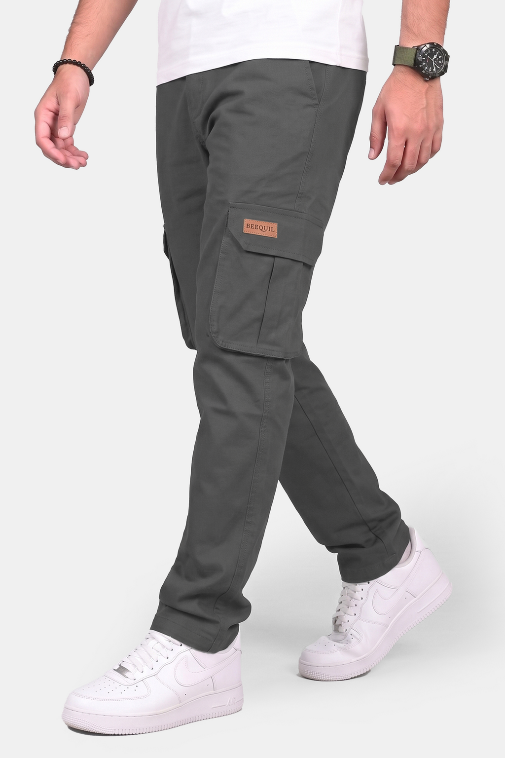 A person wearing dark gray cargo pants with a brown patch on the side pocket, styled with a white t-shirt and white sneakers against a light gray background.