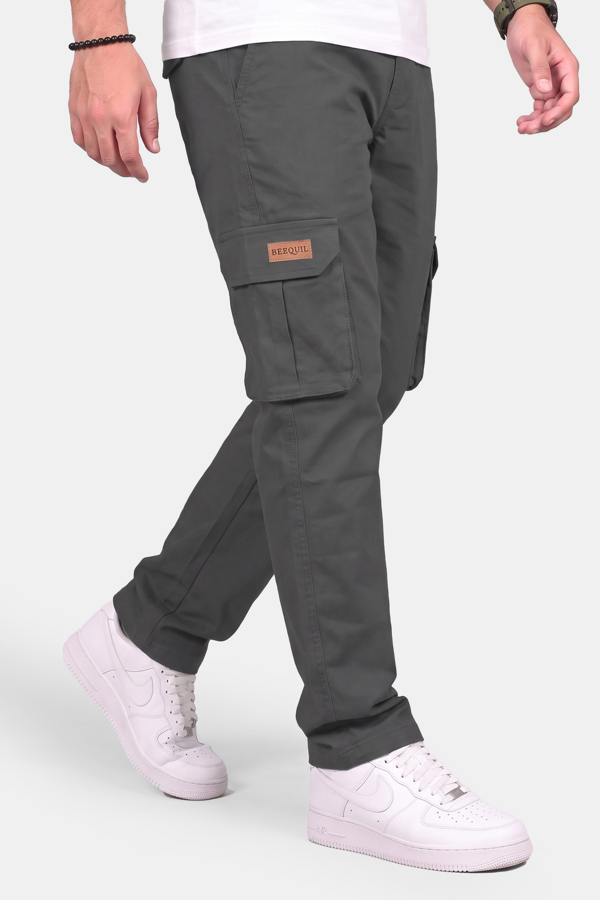 A person wearing dark gray cargo pants with a tan brand patch on the side pocket, paired with white sneakers, shown from the waist down against a light gray studio background.