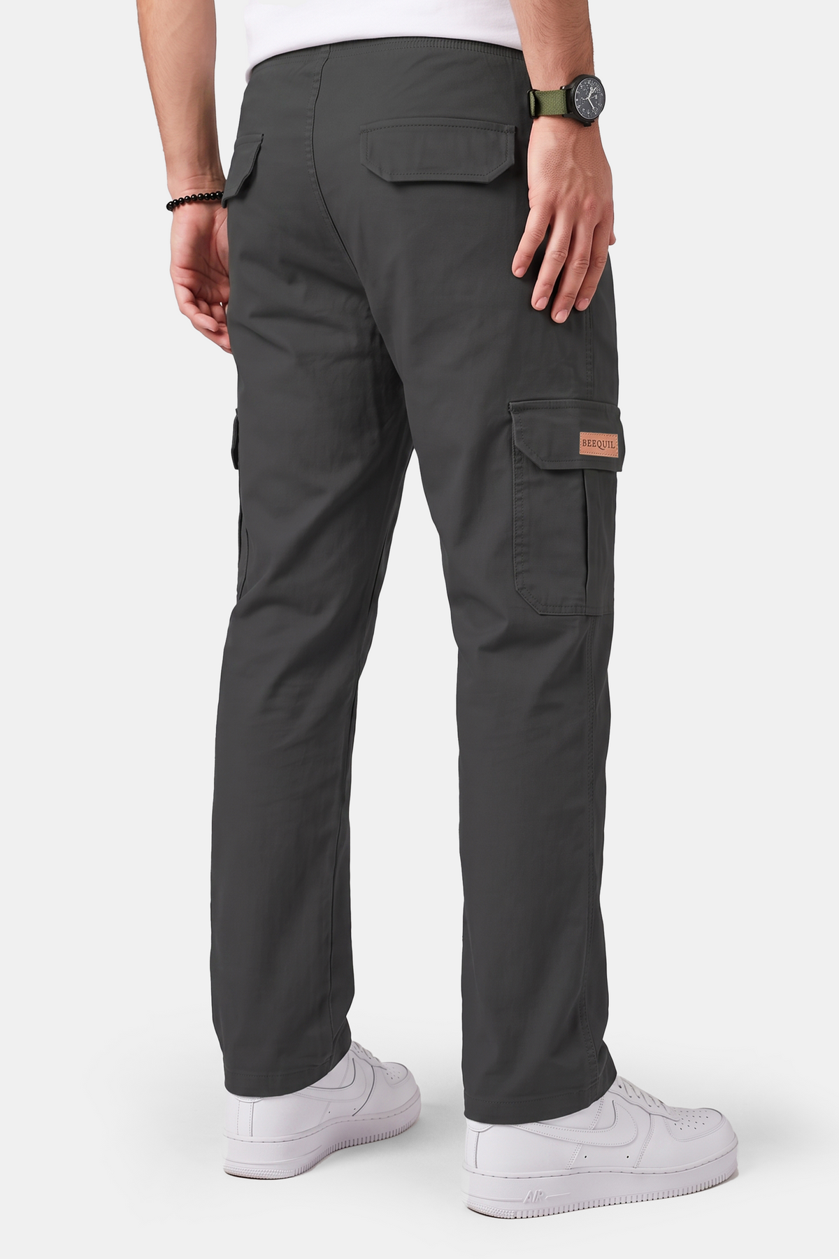 Rear view of a person wearing dark charcoal cargo pants with white sneakers and a black watch, shown against a light gray studio background.