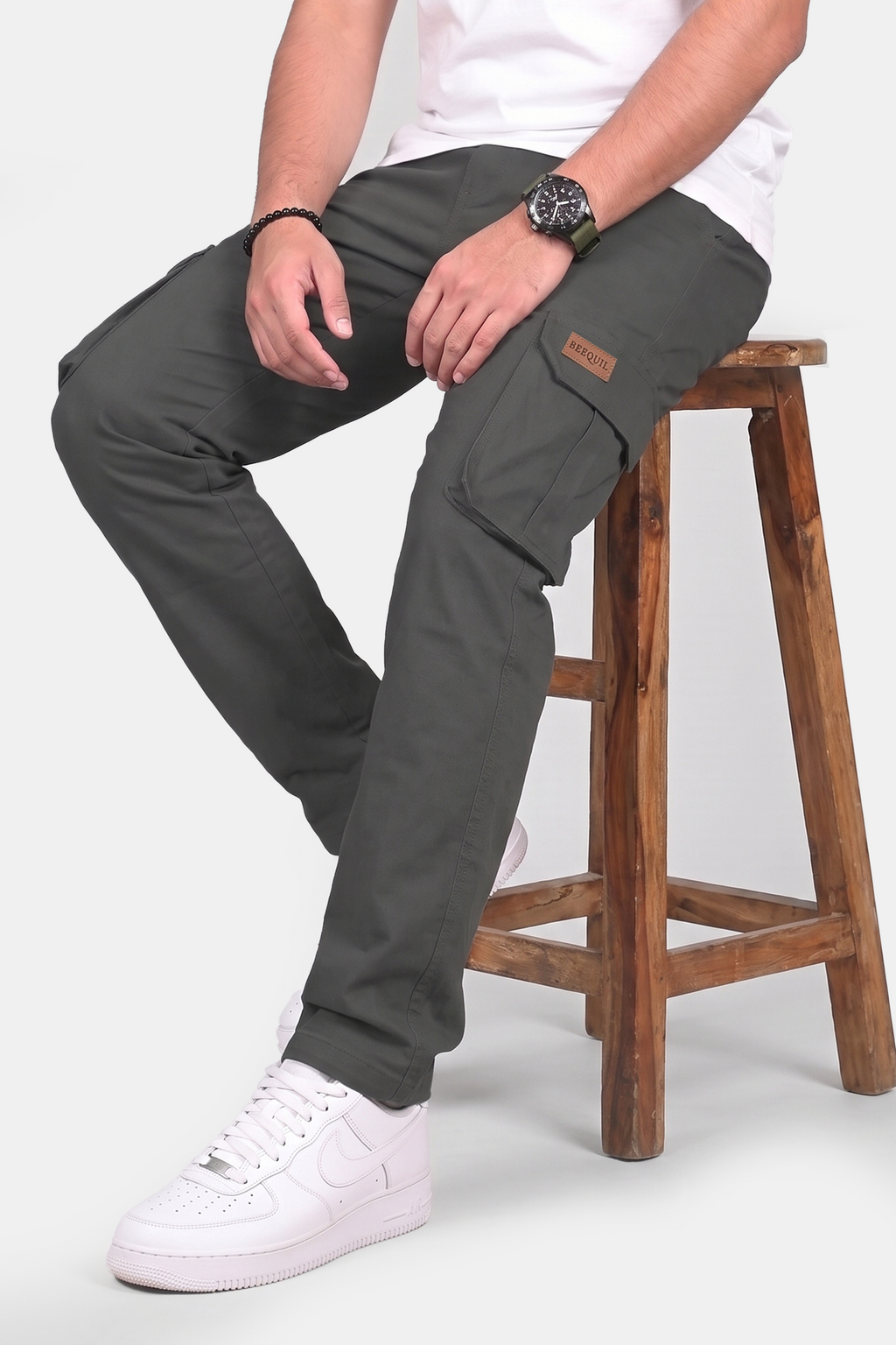 A man wearing dark gray cargo pants sits on a wooden stool, paired with a white t-shirt and white sneakers. The photo is taken from the waist down against a light gray studio background.