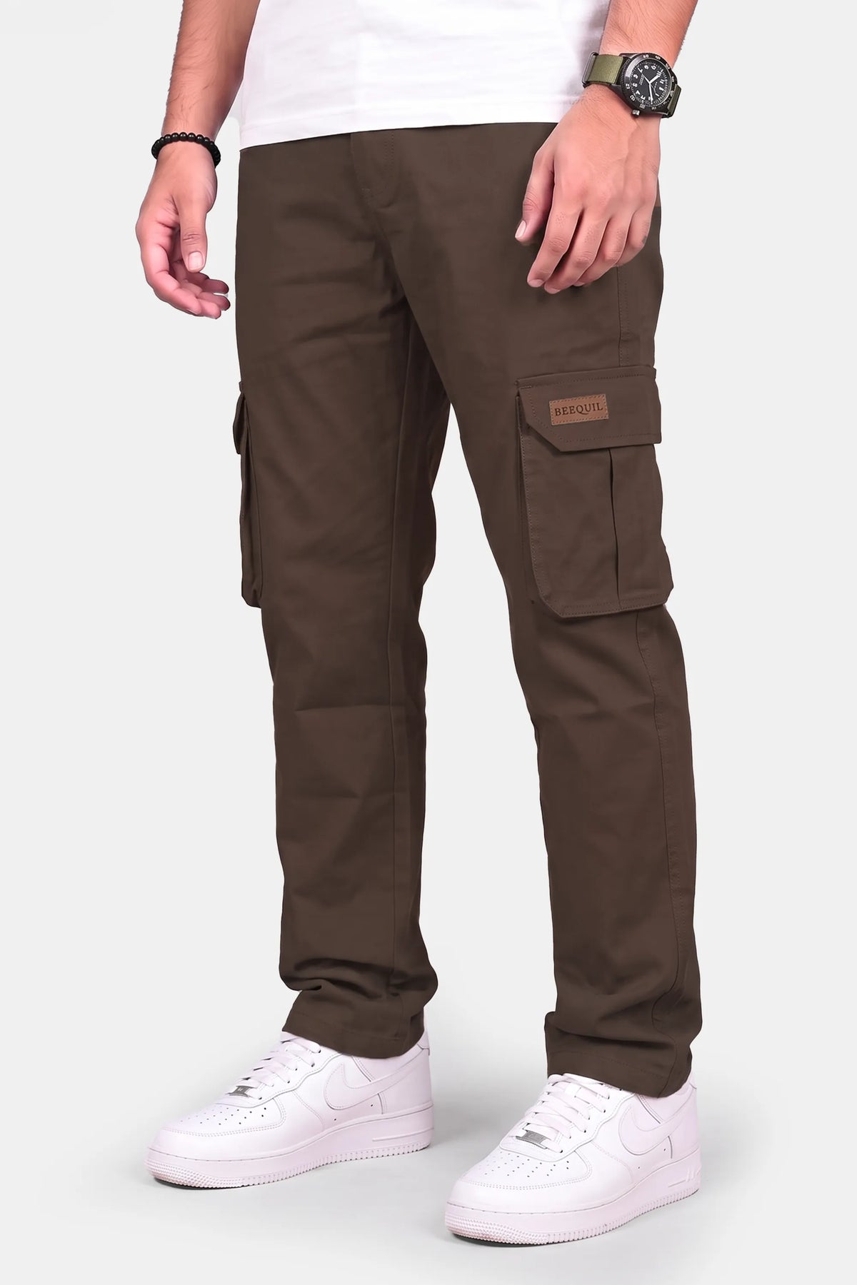 A person wearing dark brown cargo pants with side flap pockets, styled with a white t-shirt and white sneakers against a light gray studio background.