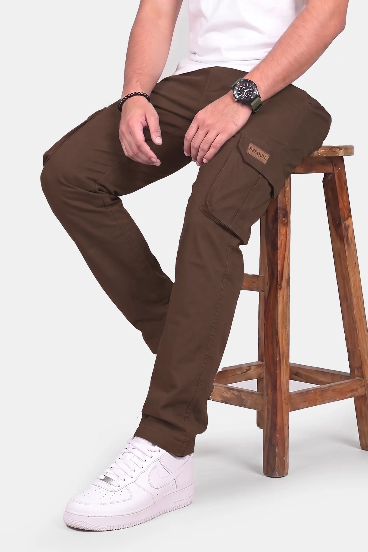 A man wearing brown cargo pants sits on a wooden stool, showing off the side pockets and fit. He is also wearing a white t-shirt and white sneakers against a light gray background.