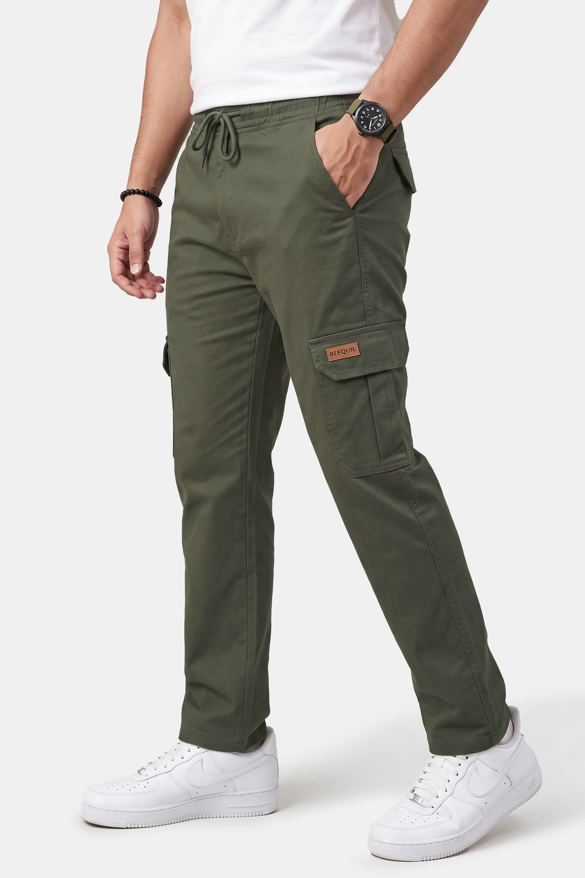 A man wearing olive green cargo pants with a drawstring waist, side cargo pocket, and a small orange label, paired with a white t-shirt and white sneakers against a light gray studio background.