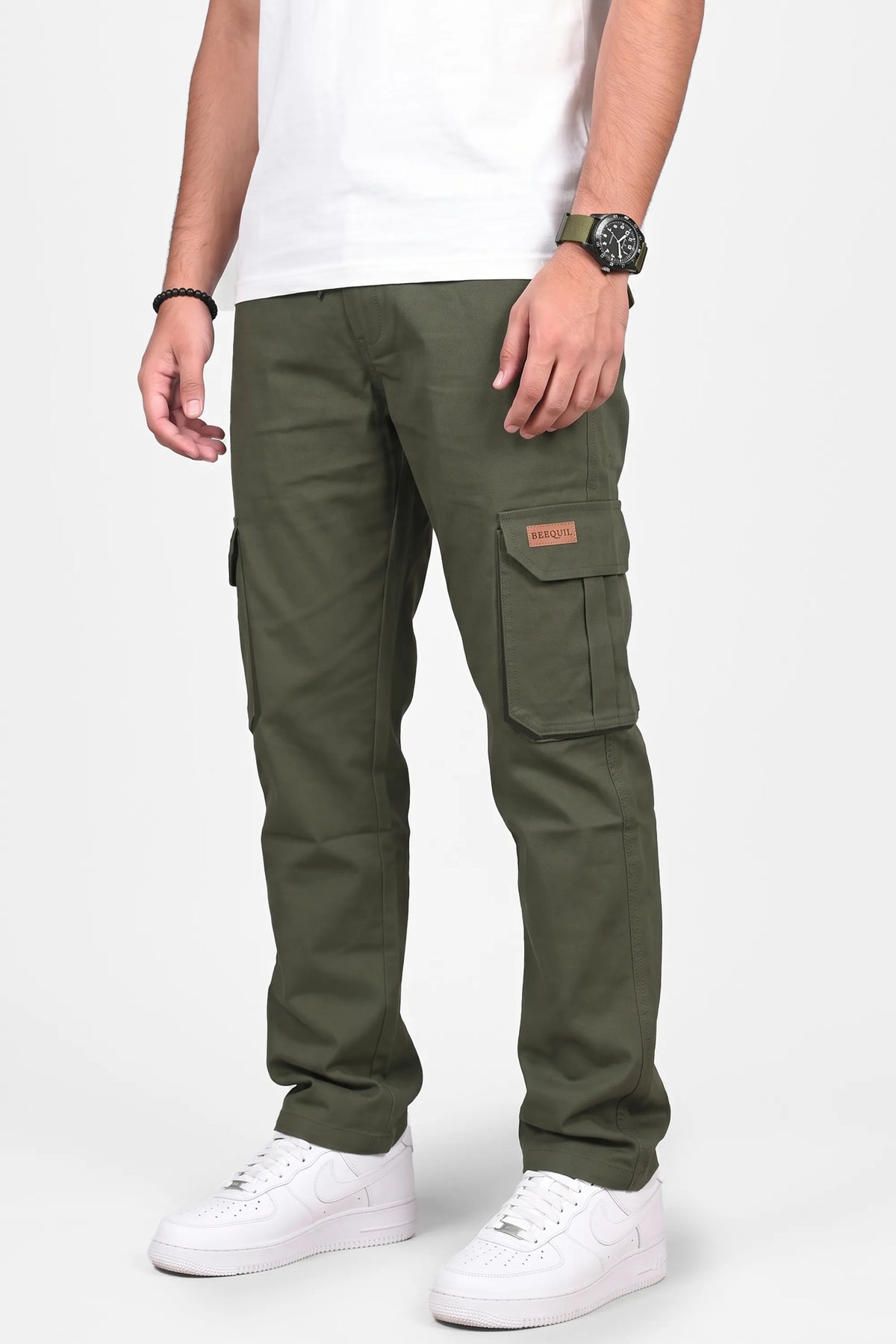 A person wearing olive green cargo pants with side flap pockets, paired with a white t-shirt and white sneakers, shown from the waist down on a light gray studio background.